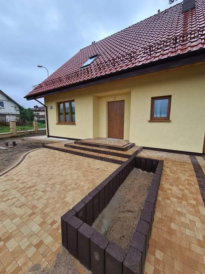 Ułożona kostka brukowa w odcieniach beżu i brązu prowadząca do domu w Pruszczu Gdańskim, z obrzeżem z ciemnej kostki tworzącym rabatę na rośliny.
