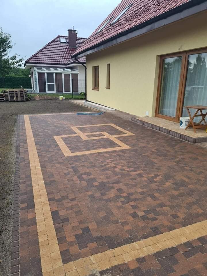 Ułożona dwukolorowa kostka brukowa z geometrycznym wzorem na podjeździe przed domem z beżową elewacją i brązowymi oknami, widoczne obrzeża z ciemnej kostki oraz przygotowany teren pod trawnik.