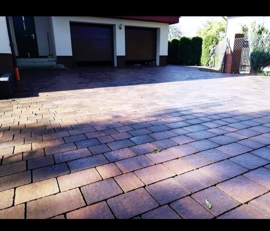 Ułożona, brązowo-szara kostka brukowa przed budynkiem z dwoma bramami garażowymi, widoczne pojedyncze liście na nawierzchni.