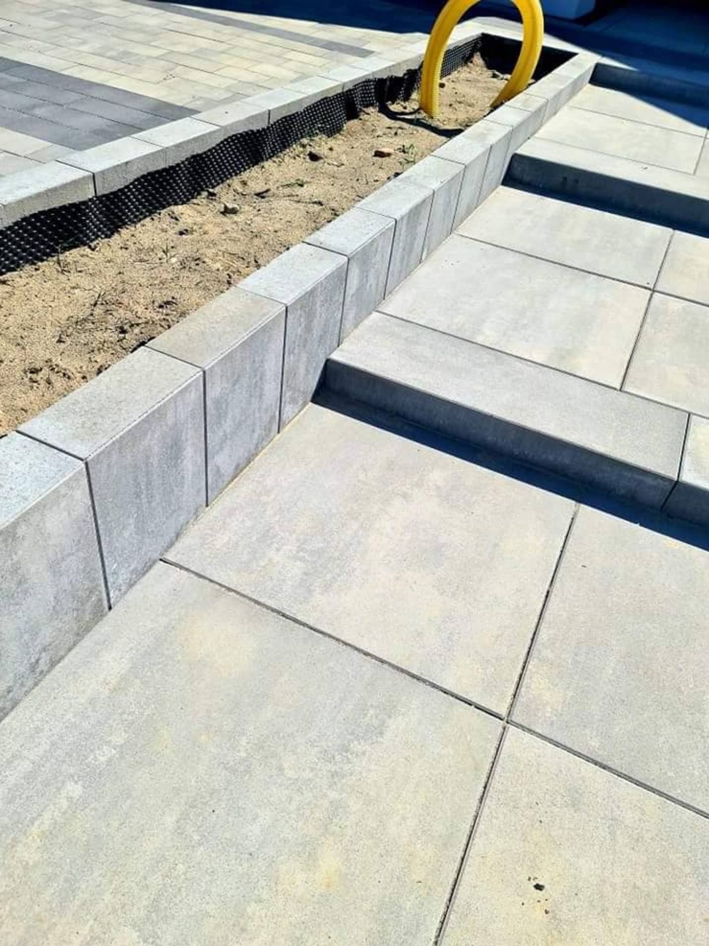 Szara nawierzchnia z płyt betonowych, obrzeże z kostki i rabata z żółtą rurą drenażową w tle.