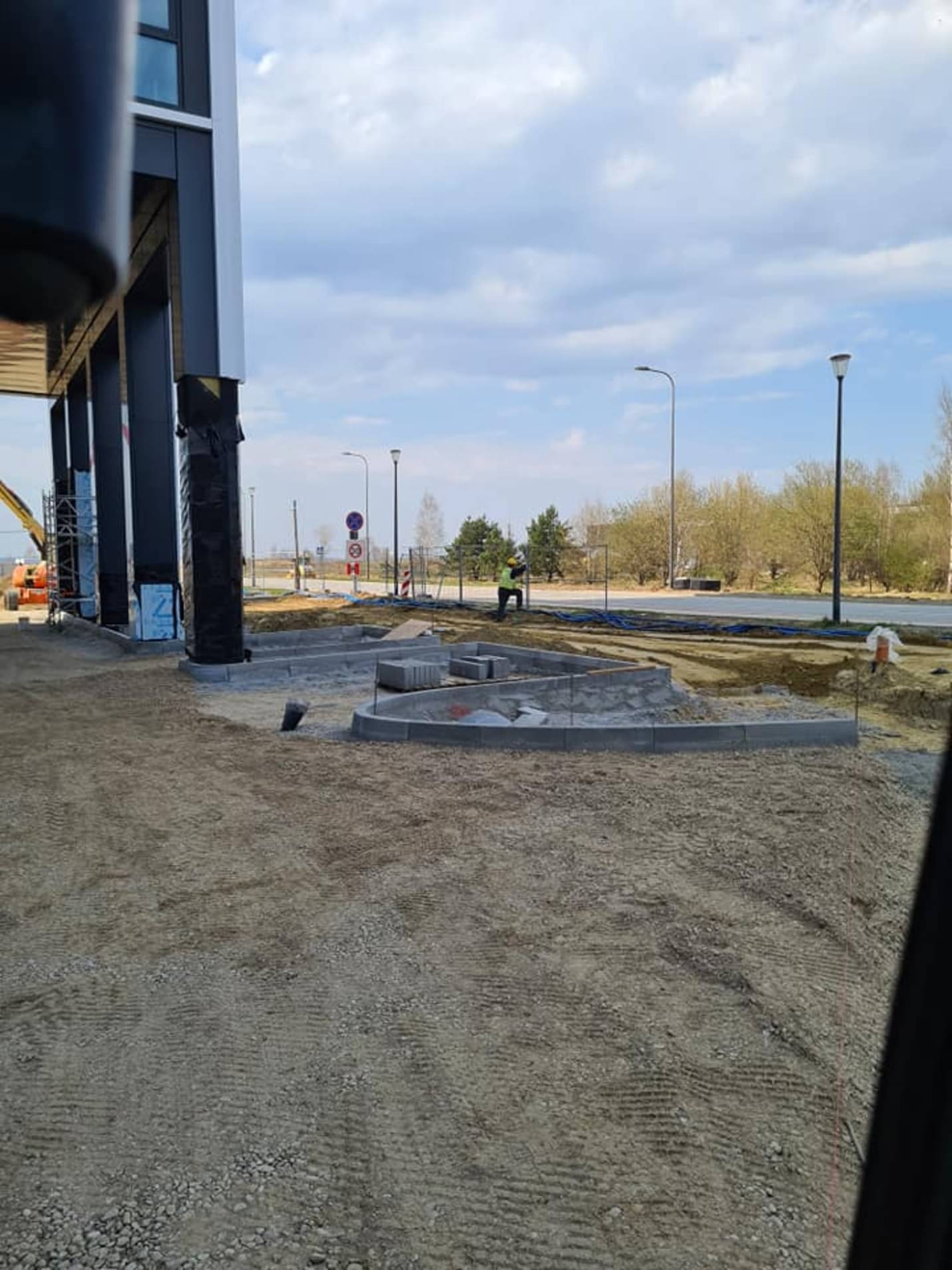 Montaż obrzeży betonowych na przygotowanym podłożu, widoczny fragment budynku z kolumnami, w tle pracownik w żółtej kamizelce i maszyna budowlana.