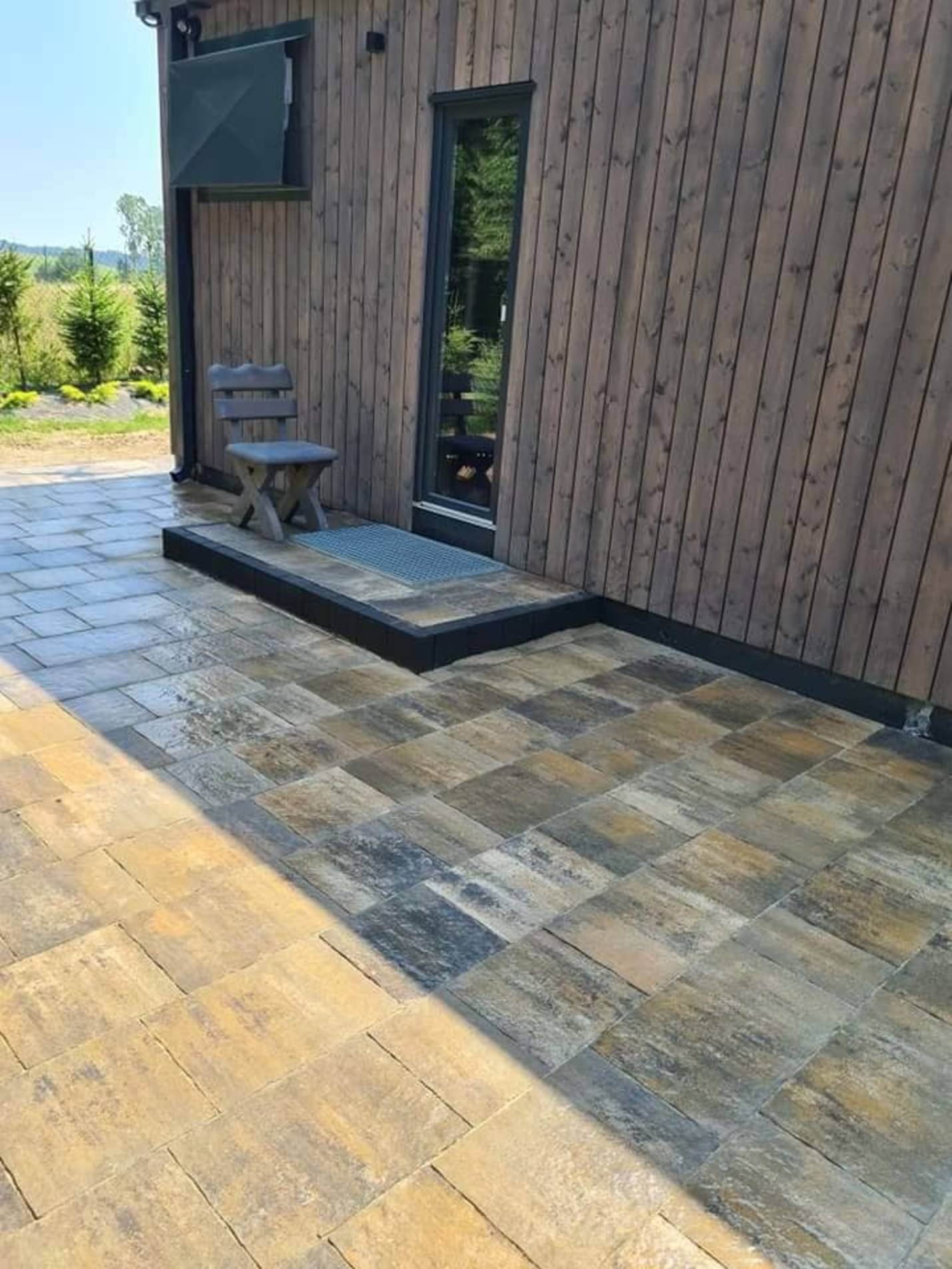 Nowoczesny taras z kostki brukowej w odcieniach beżu i szarości, przylegający do drewnianego domu z ciemnymi panelami, z prostym drewnianym krzesłem i wycieraczką przed wejściem.