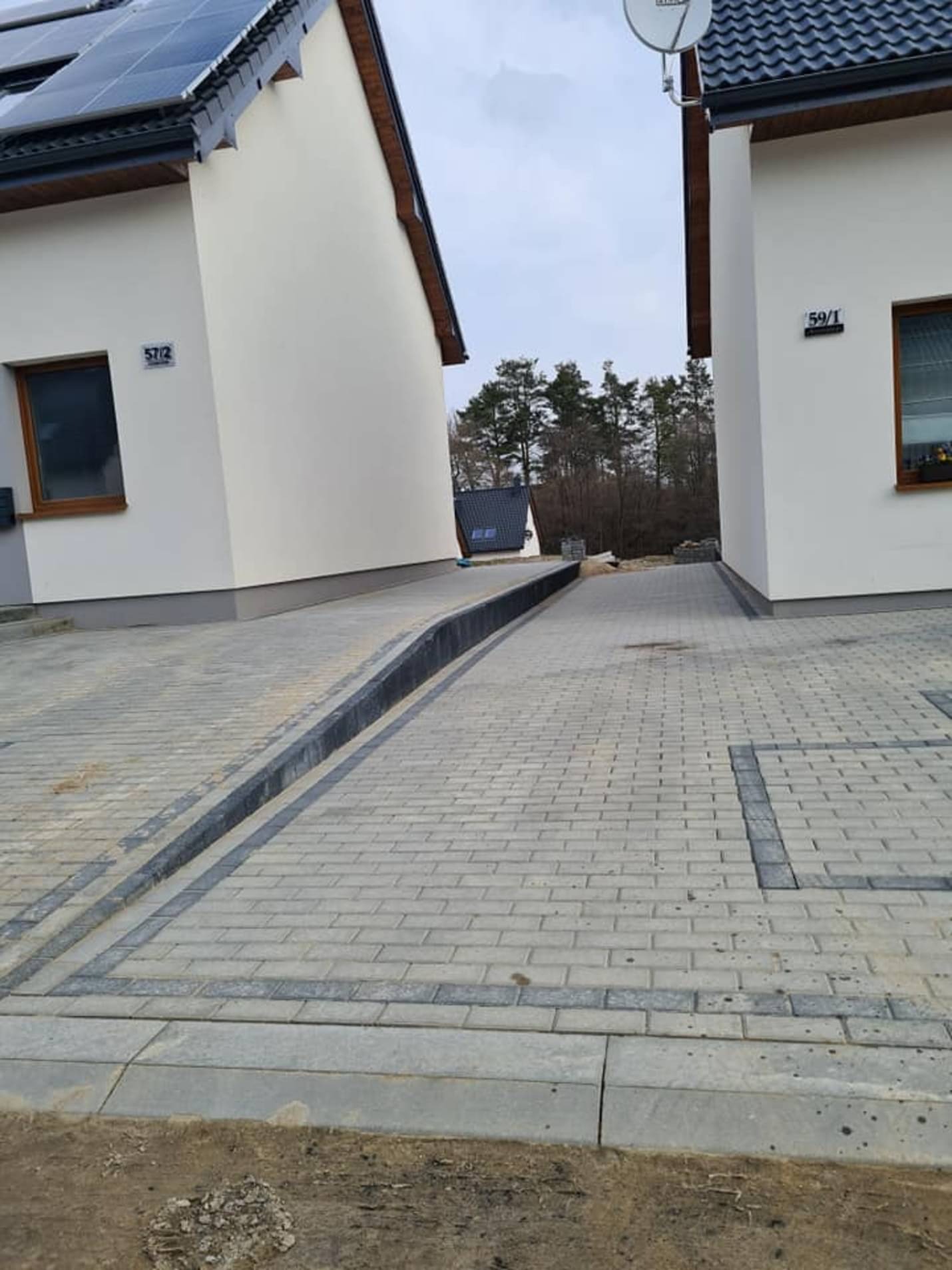 Ułożona szara kostka brukowa przed dwoma nowoczesnymi domami z panelami słonecznymi, z widocznym odpływem liniowym i obrzeżami.