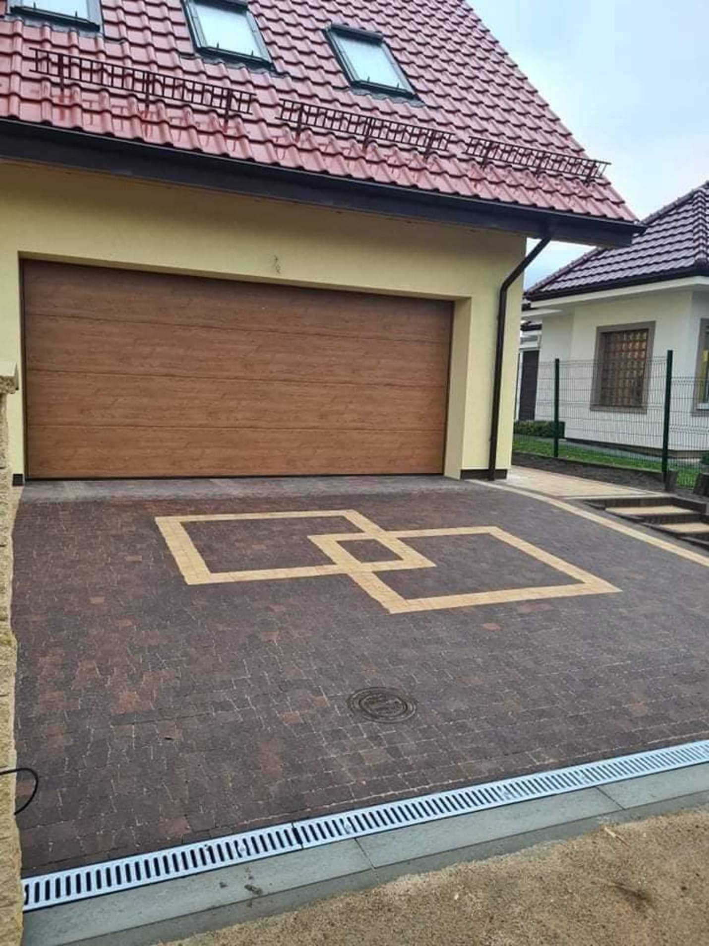 Podjazd z kostki brukowej w odcieniach brązu z geometrycznym wzorem w kolorze beżowym, prowadzący do garażu z brązową bramą segmentową, obok widoczny budynek mieszkalny z czerwoną dachówką.