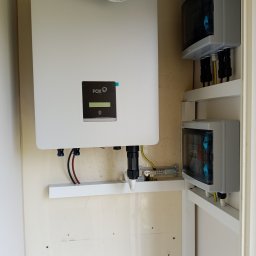 Samowystarczalni.eu Sp. z o.o. - Biały inwerter Fox ESS z podłączeniami kablowymi i dwoma szarymi rozdzielnicami wewnątrz szafki, widoczne przewody i listwa zaciskowa.