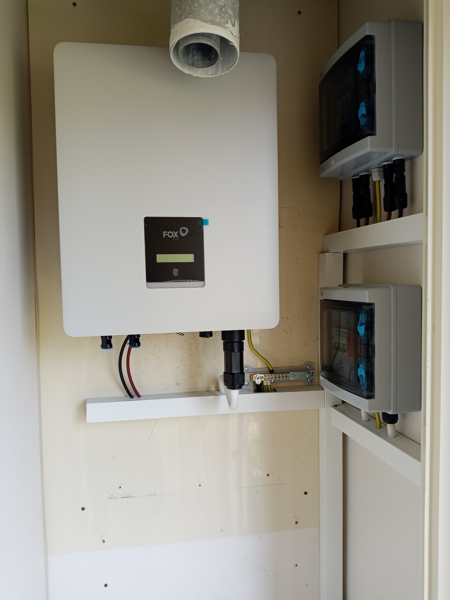 Biały inwerter Fox ESS z podłączeniami kablowymi i dwoma szarymi rozdzielnicami wewnątrz szafki, widoczne przewody i listwa zaciskowa.