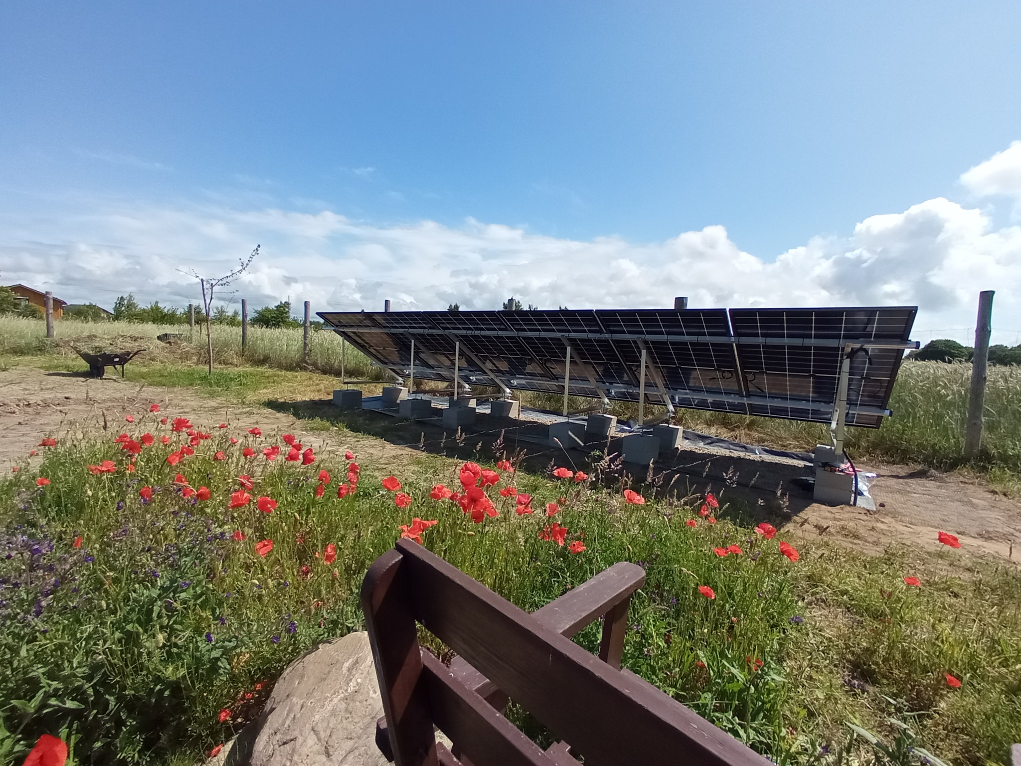 Instalacja paneli fotowoltaicznych na łące z polnymi kwiatami, widok zza drewnianej ławki. Panele zamontowane na betonowych podstawach, błękitne niebo z chmurami w tle.