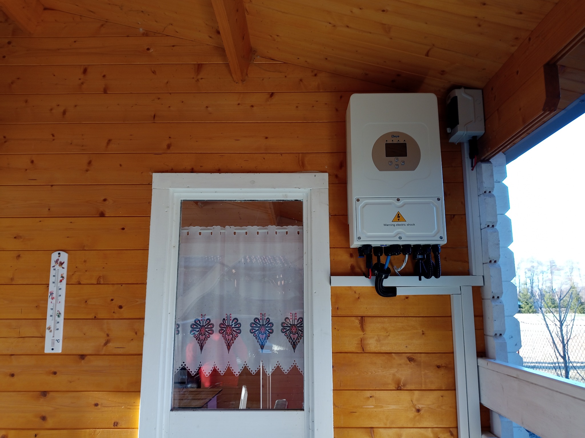 Biały inwerter fotowoltaiczny marki Deye zamontowany na drewnianej ścianie domku, obok okna z firanką i termometrem. Widoczne kable i elementy montażowe.