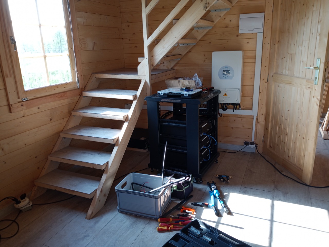 Wnętrze drewnianego domku z modułami energii na metalowym stojaku, narzędziami i schodami. Widoczny inwerter fotowoltaiczny na ścianie.