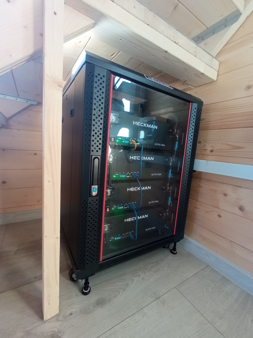 Czarna szafa rack Heckman z zainstalowanymi urządzeniami, umieszczona w drewnianym pomieszczeniu z jasną podłogą, widok z boku.