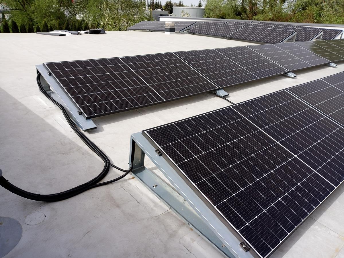 Rząd paneli fotowoltaicznych na płaskim dachu, zamontowanych na metalowych konstrukcjach. Widoczne kable instalacyjne. Ujęcie z góry.