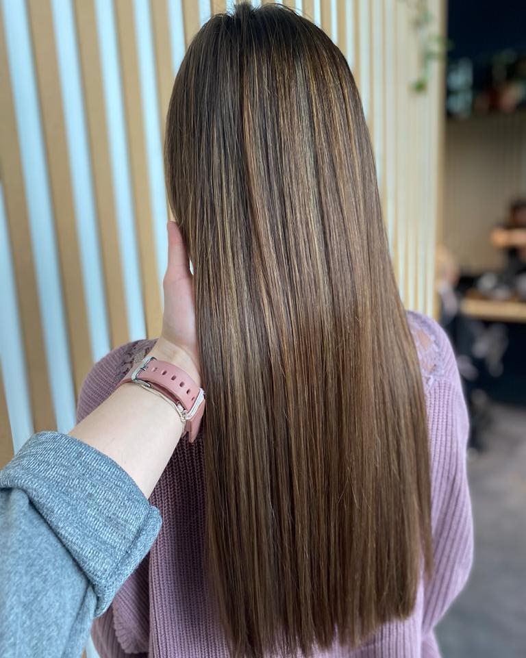 Długie, proste włosy w odcieniach brązu z refleksami, widok z tyłu, dłoń z różowym smartwatchem dotyka włosów, w tle jasne pionowe panele.