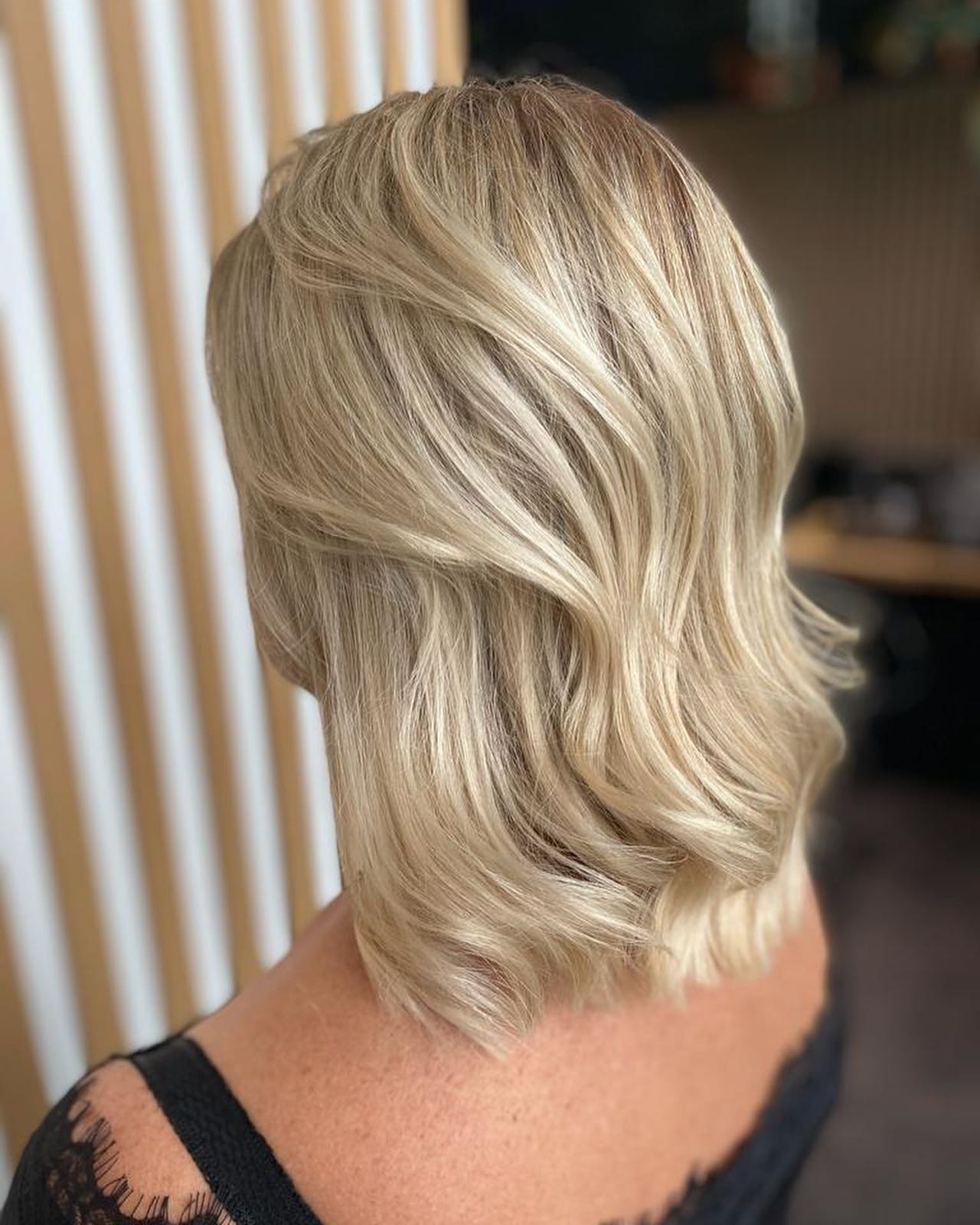 Krótkie, falowane włosy w odcieniach blondu, z delikatnymi refleksami, widoczne od tyłu, z koronkowym wykończeniem bluzki.