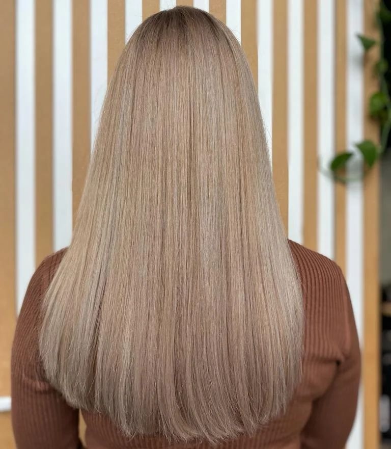 Długie, proste włosy w odcieniu chłodnego blondu, widok z tyłu, na tle ściany z pionowymi paskami i rośliną doniczkową.
