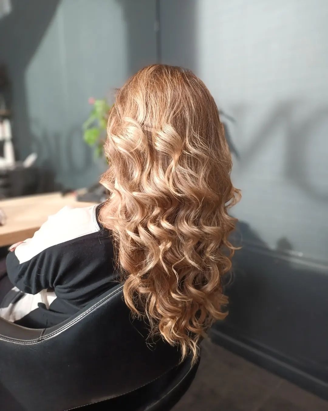 Długie, blond włosy z trwałymi lokami, widok z tyłu, siedząca osoba w czarno-białej bluzie na czarnym fotelu w salonie.