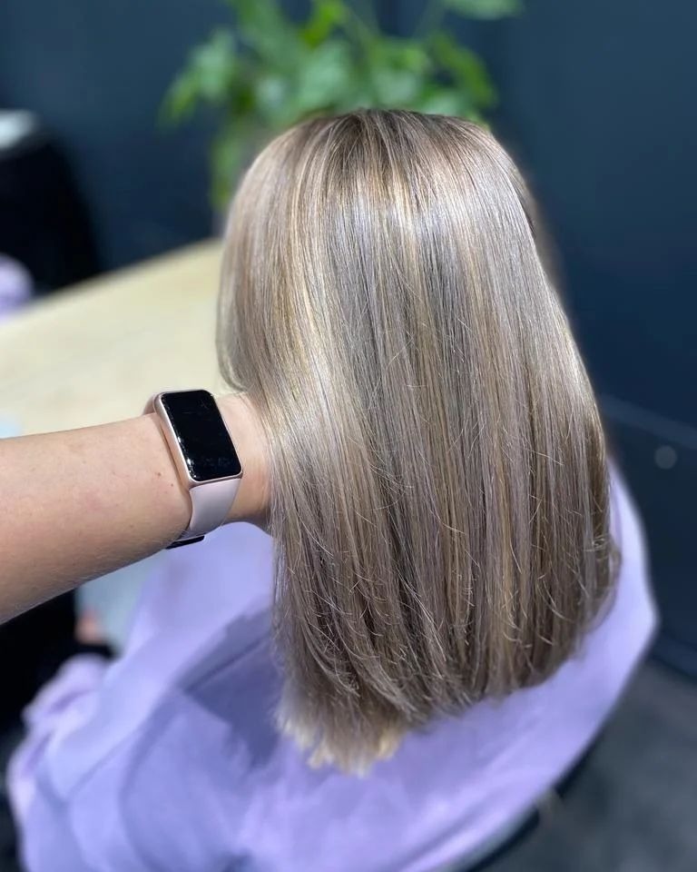 Włosy blond z refleksami po koloryzacji, widoczne od tyłu, ręka z zegarkiem w tle, stonowane oświetlenie.