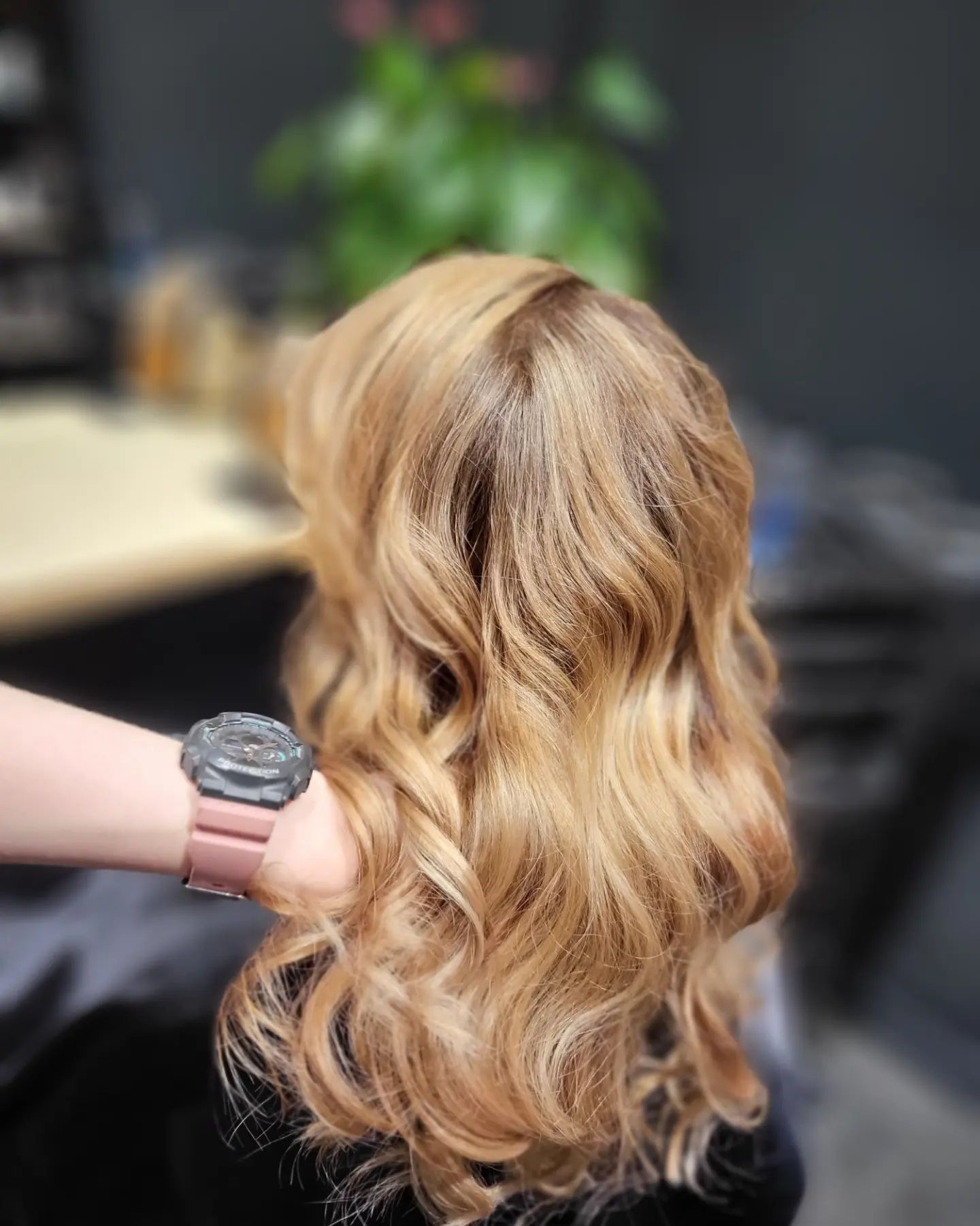 Tył głowy z blond włosami ułożonymi w fale, trzymanymi przez rękę z zegarkiem na brzoskwiniowym pasku, rozmyte tło salonu.