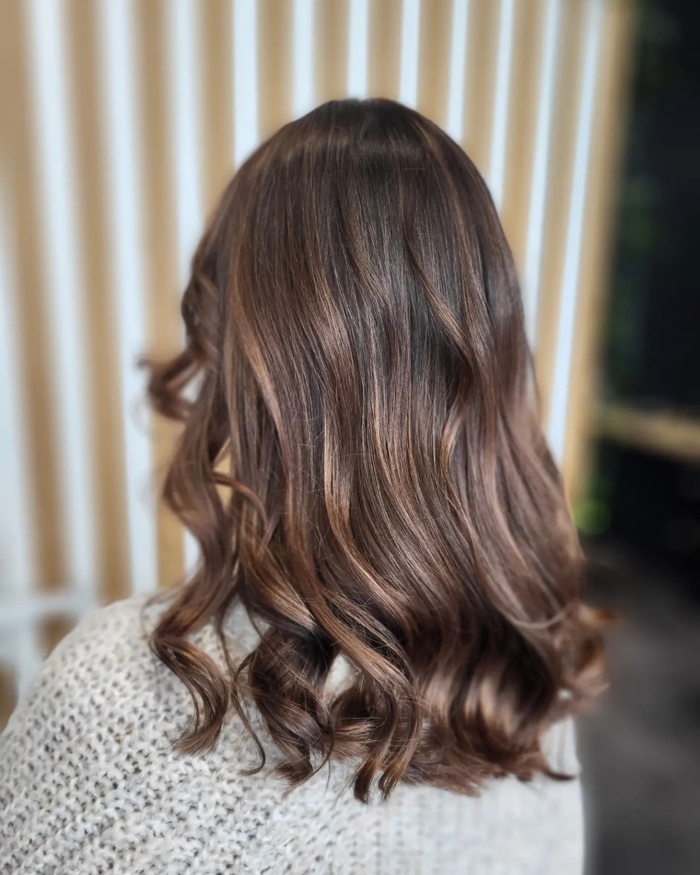 Ciemnobrązowe włosy z subtelnymi refleksami, delikatnie pofalowane na końcach, widoczne z tyłu, na tle pionowych, kremowych paneli.