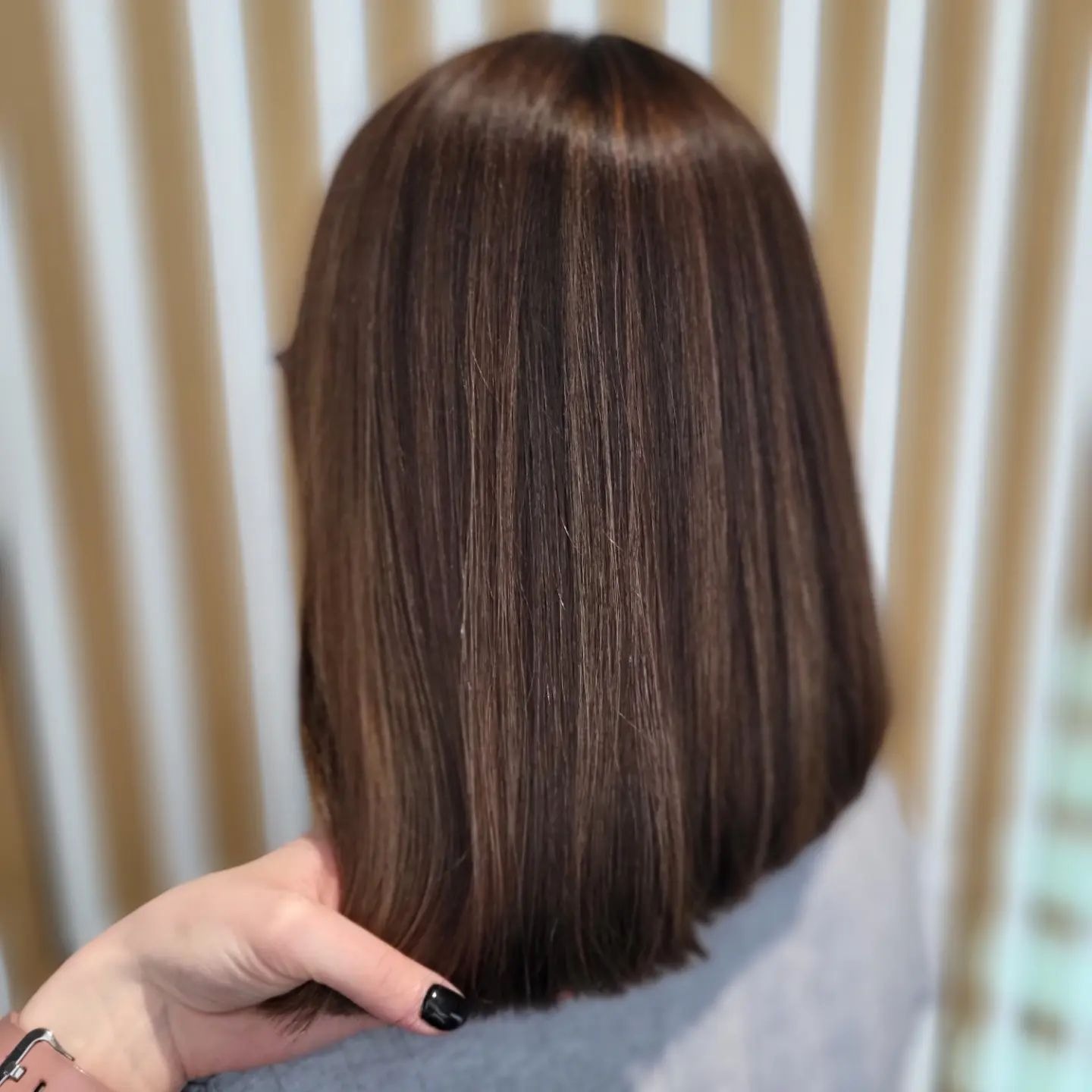 Brązowe włosy do ramion z delikatnymi refleksami, widok z tyłu, dłoń kobiety z czarnym manicure przytrzymuje końcówki włosów.