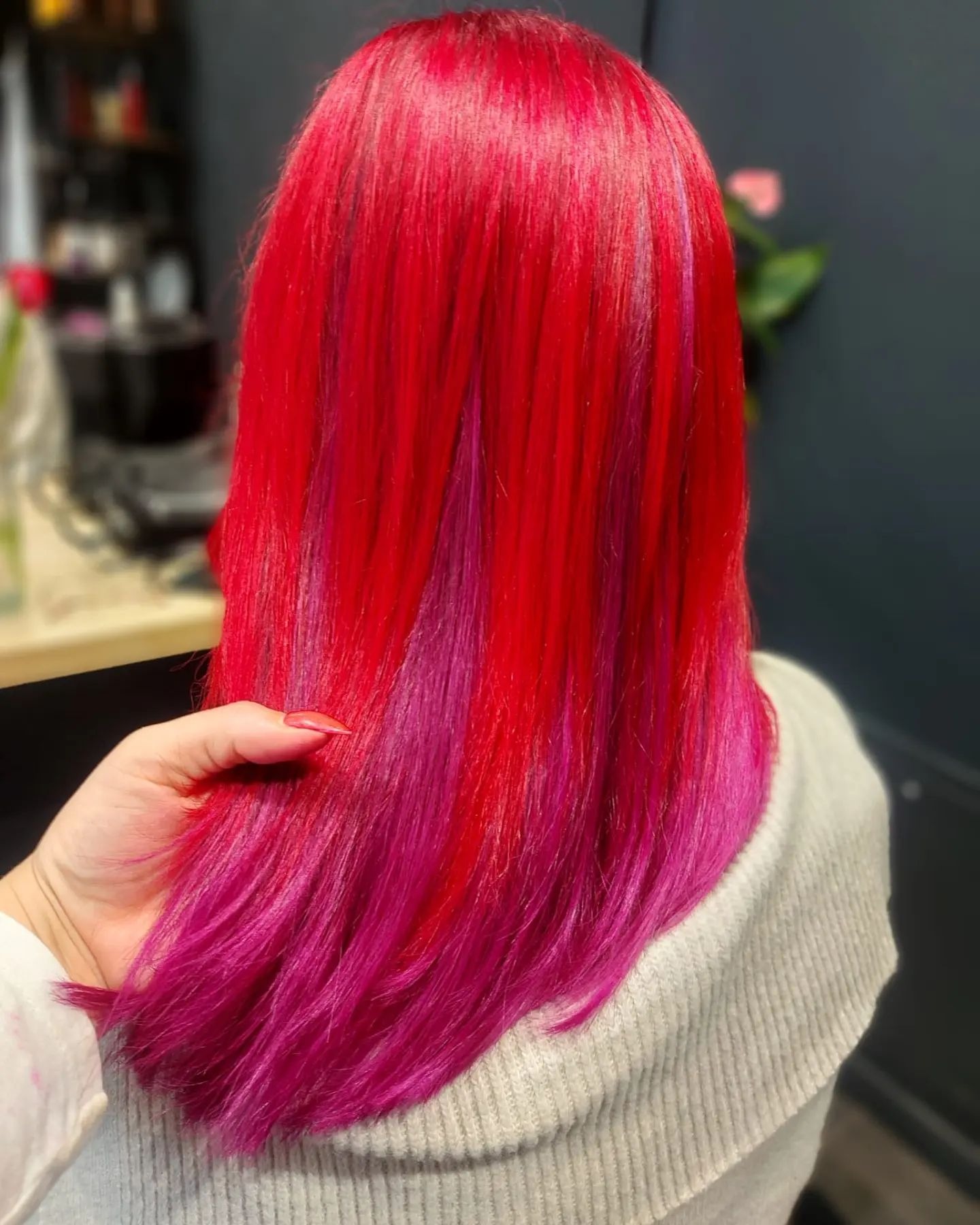 Intensywnie czerwone włosy z różowymi pasemkami, widoczne od tyłu, stylizowane w Bytomiu. Dłoń z czerwonym manicure delikatnie unosi kosmyki. Tło rozmyte, widoczne elementy wyposażenia salonu.