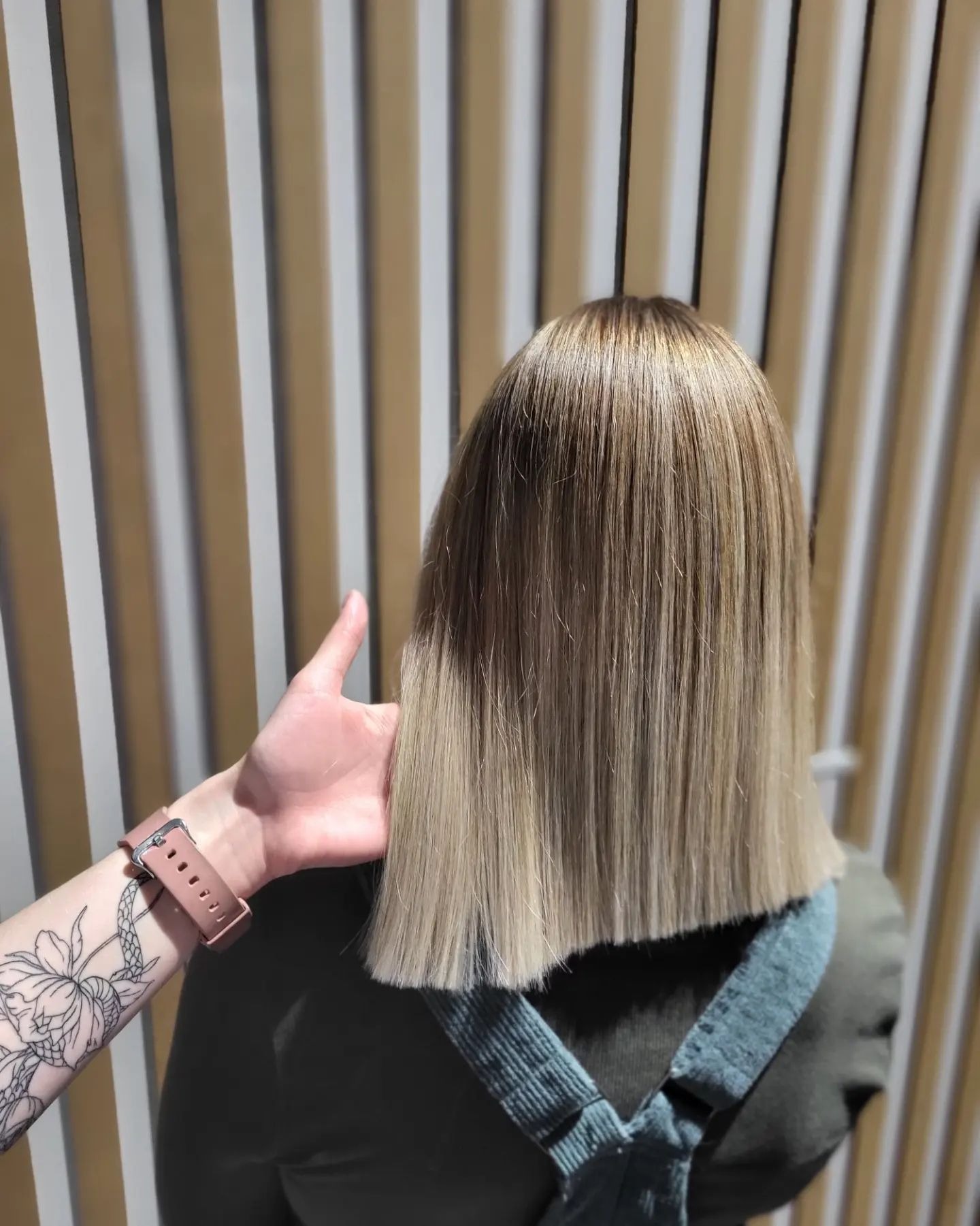 Proste, blond włosy do ramion z ciemniejszym odrostem, widok z tyłu, dłoń z tatuażem kwiatowym przytrzymująca włosy, tło z pionowych pasków.