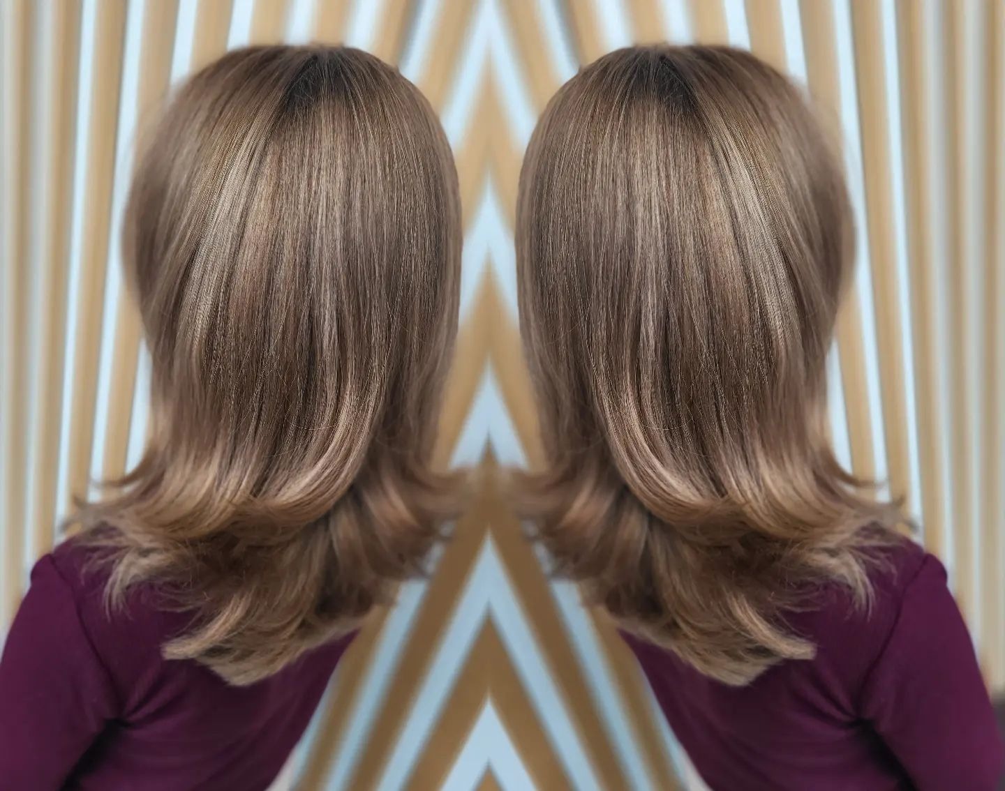 Damska fryzura warstwowa z refleksami, widok z tyłu na tle ściany z geometrycznym wzorem w odcieniach beżu i bieli.