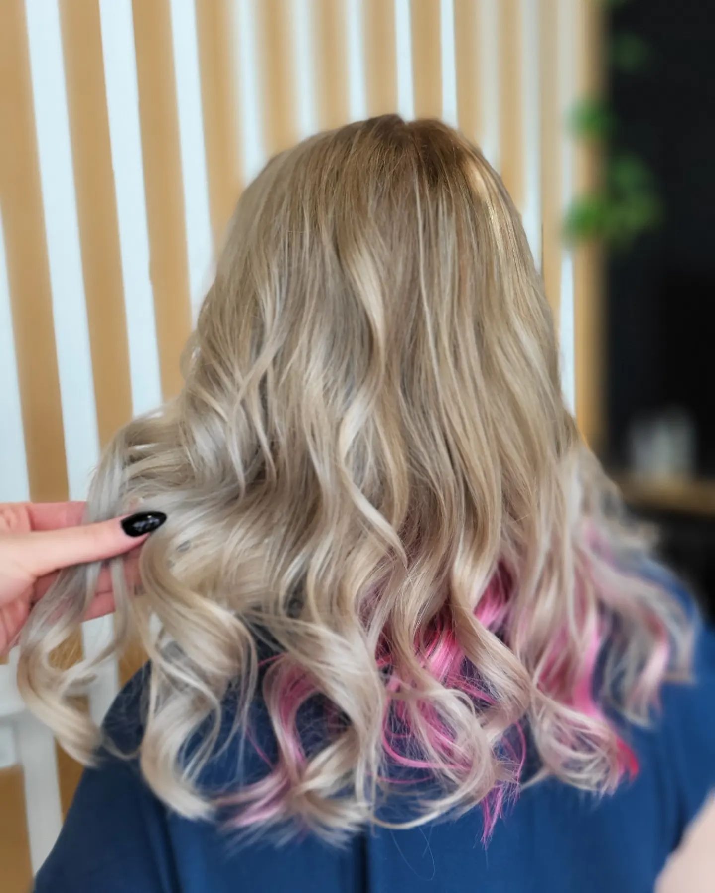 Blond włosy z różowymi pasemkami u nasady, delikatne fale, dłoń z czarnym manicure poprawia kosmyk, tło z pionowych, jasnych paneli.