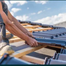 Dekarz układa rząd ciemnoszarych dachówek na drewnianej konstrukcji dachu, widoczne niebo z lekkimi chmurami.