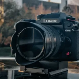 Aparat Lumix GH6 z założonym obiektywem i osłoną przeciwsłoneczną, umieszczony na precyzyjnej szynie montażowej, z rozmytym tłem miejskim.