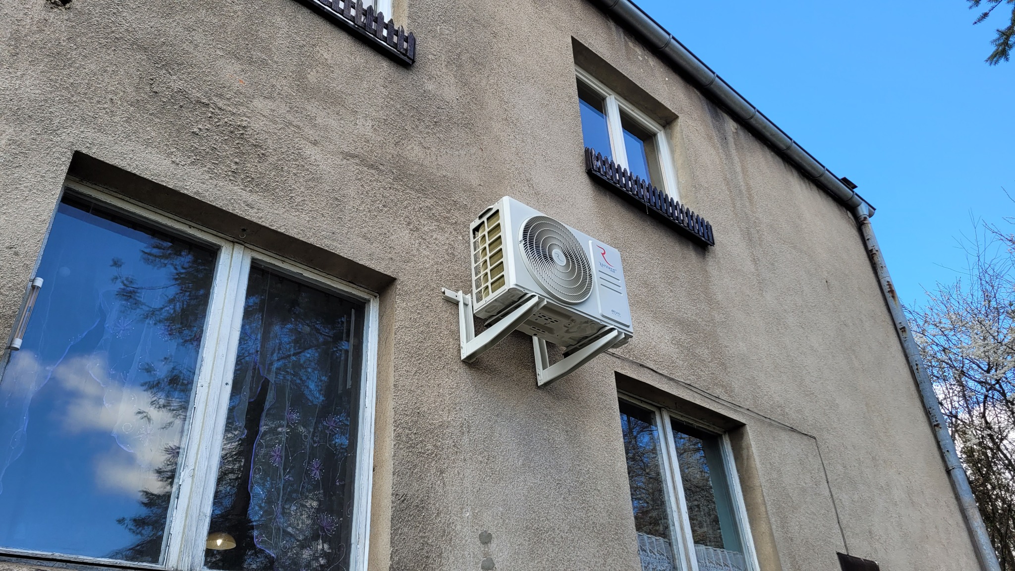 Biała jednostka zewnętrzna klimatyzatora zamontowana na szarej elewacji budynku mieszkalnego, widoczna z dołu, z oknami z drewnianą stolarką i metalowymi barierkami.