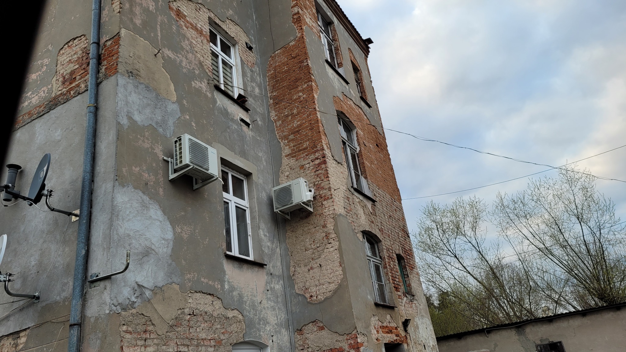 Zewnętrzne jednostki klimatyzacji zamontowane na elewacji starego, częściowo odrestaurowanego budynku z odsłoniętą cegłą i widocznymi śladami napraw.