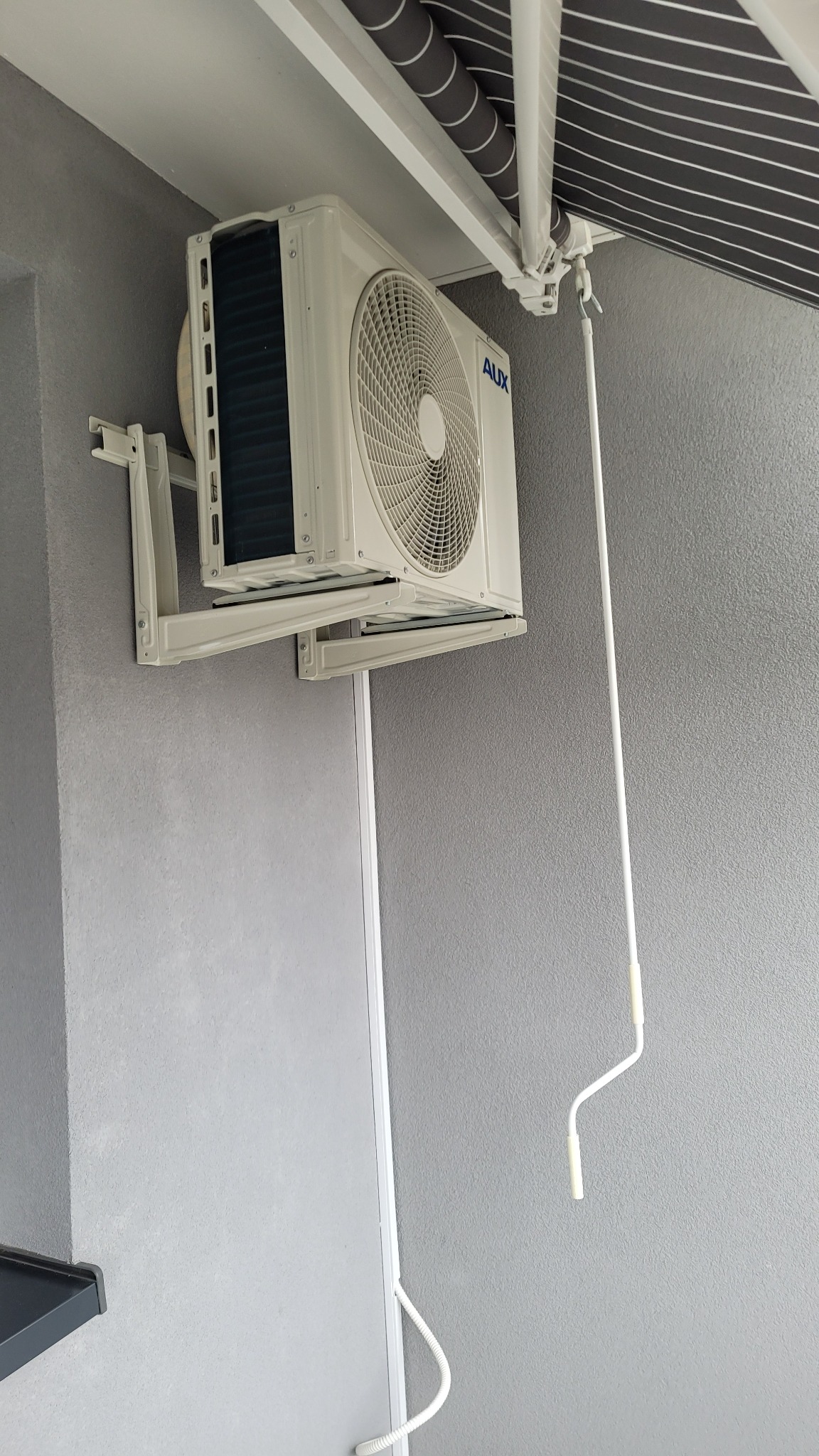 Biała jednostka zewnętrzna klimatyzacji marki AUX zamontowana na szarej ścianie pod rozwijaną markizą, widoczne wsporniki montażowe i fragment instalacji.