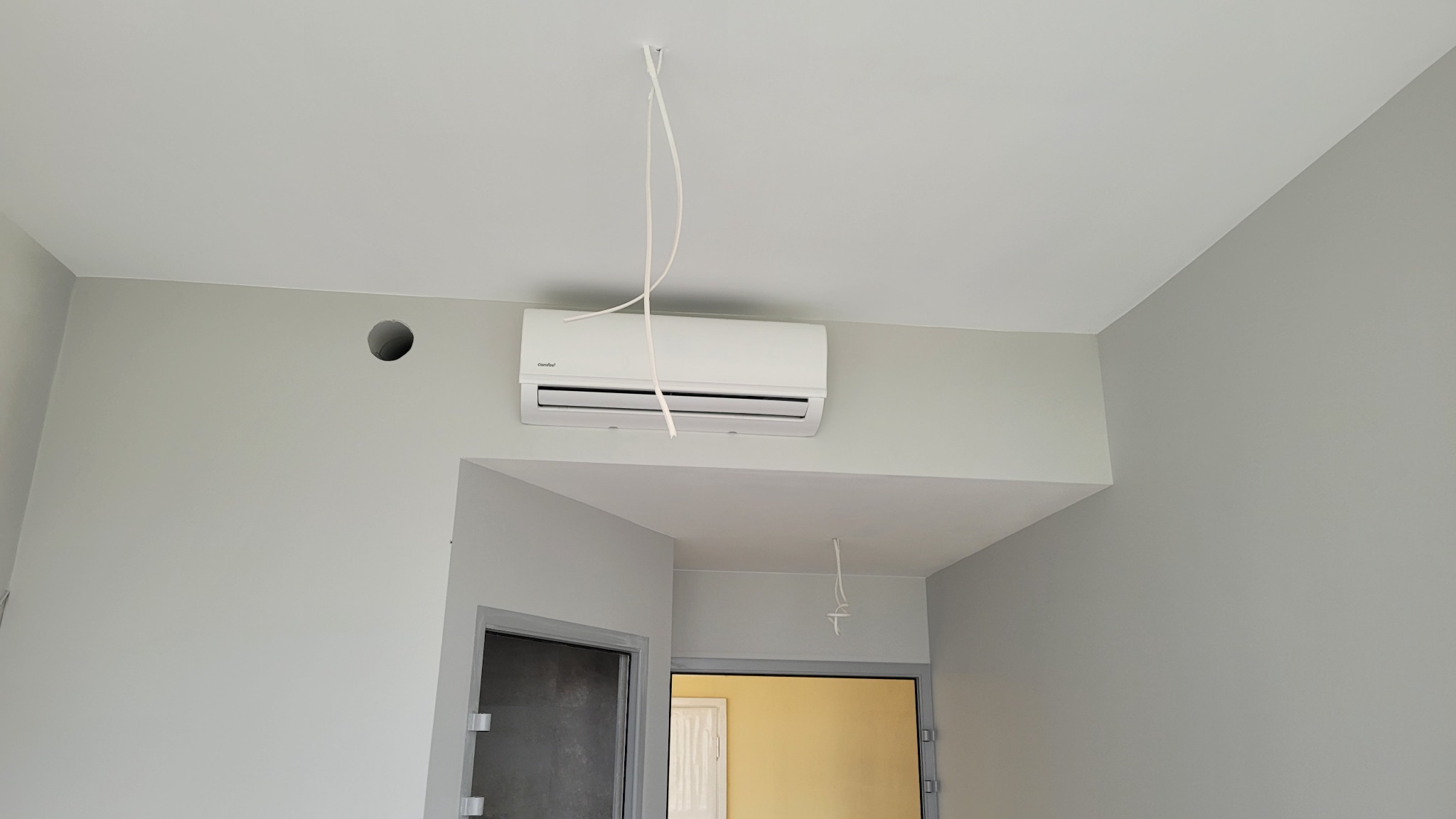 Biała jednostka klimatyzacji zamontowana na ścianie w szarym pomieszczeniu, widoczne kable zwisające z sufitu, otwór w ścianie i futryna drzwi w tle.