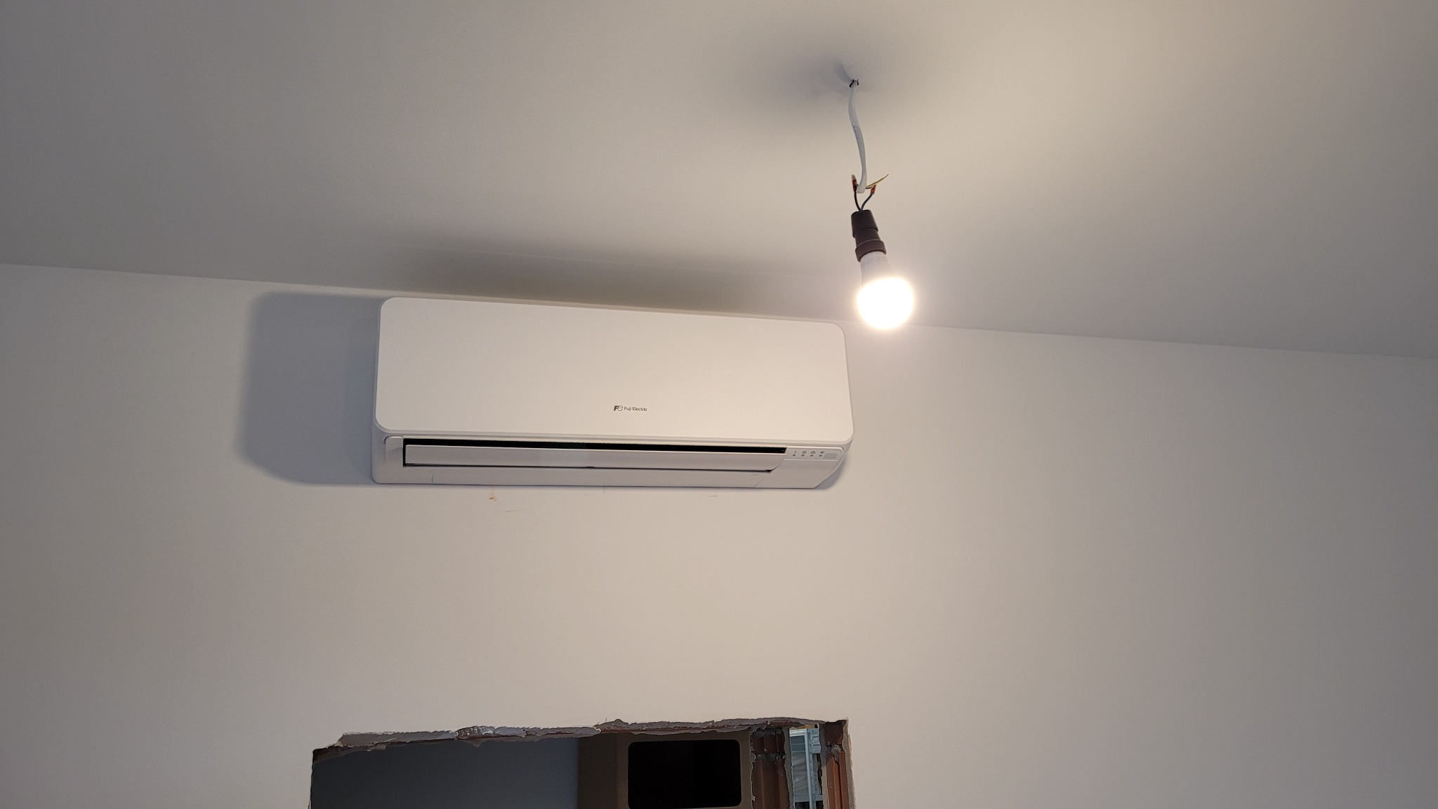 Biała jednostka klimatyzacji marki Fuji Electric zamontowana na ścianie w pomieszczeniu w trakcie remontu, widoczna prowizoryczna żarówka na kablu.