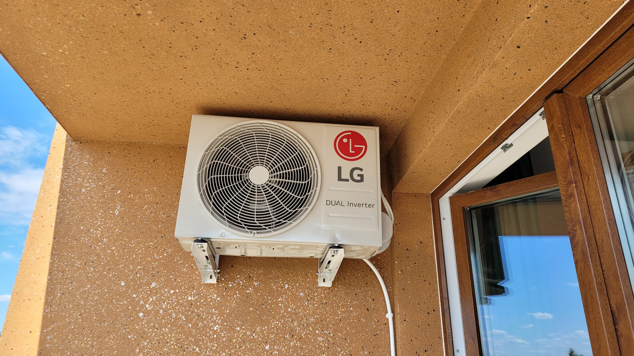 Zewnętrzna jednostka klimatyzacji LG Dual Inverter zamontowana na ścianie budynku z oknem w tle, widok od dołu.