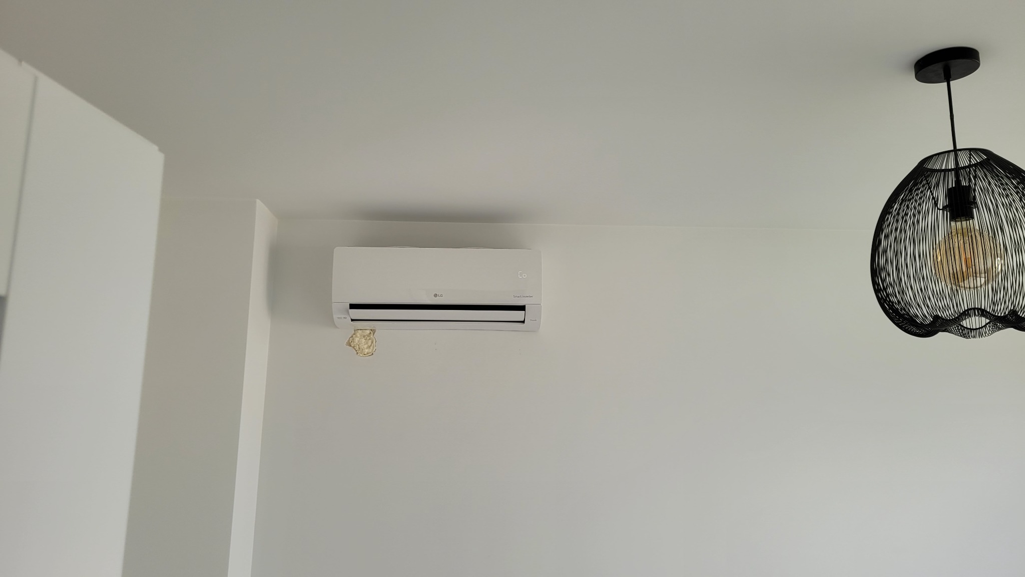 Biały klimatyzator LG Smart Inverter na białej ścianie z widoczną pianką montażową po instalacji i ozdobną lampą wiszącą z żarówką Edisona w tle.