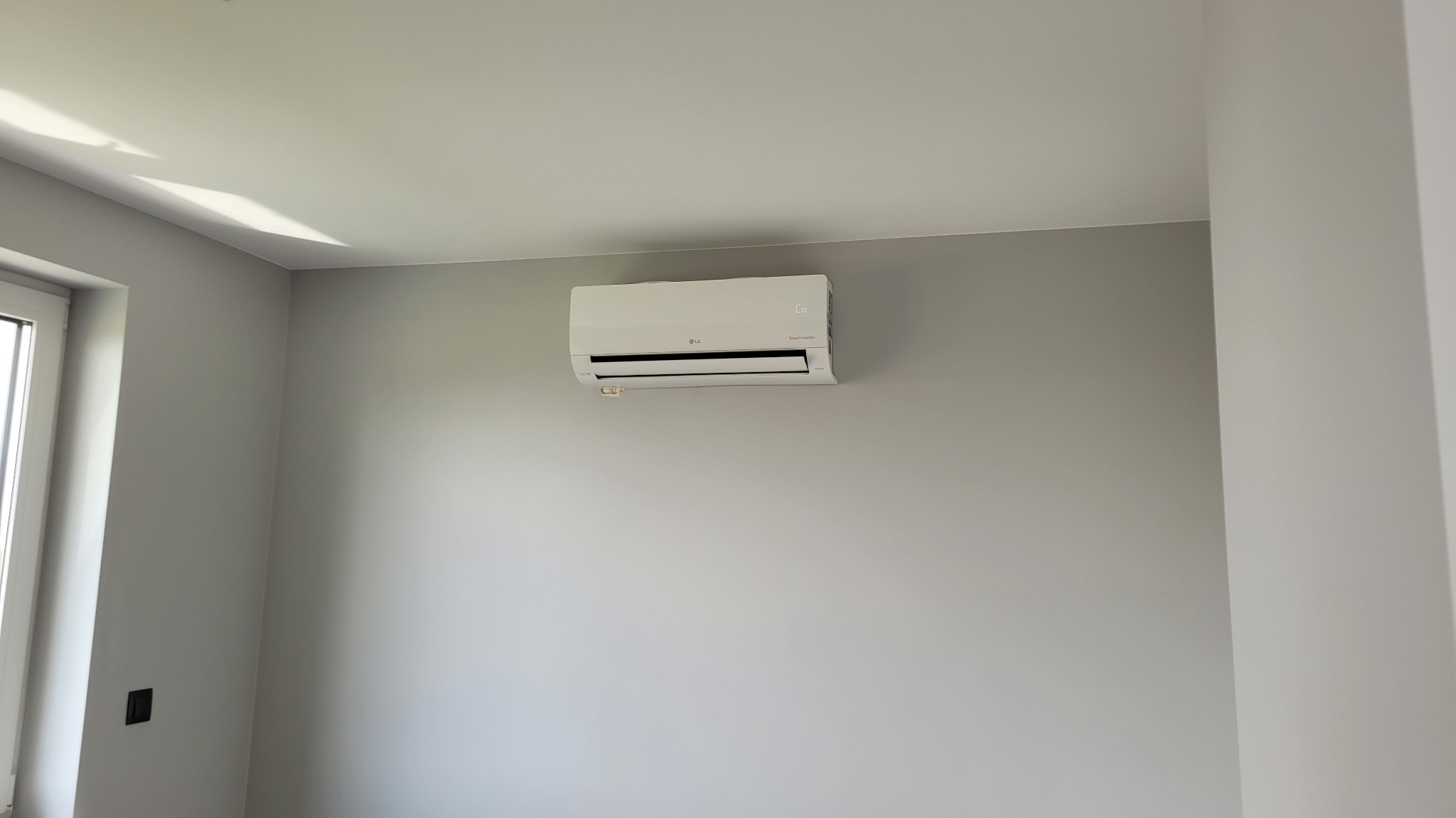 Biały klimatyzator LG Smart Inverter zamontowany na szarej ścianie w pomieszczeniu w Mąkoszycach, widok z boku, z oknem po lewej stronie.