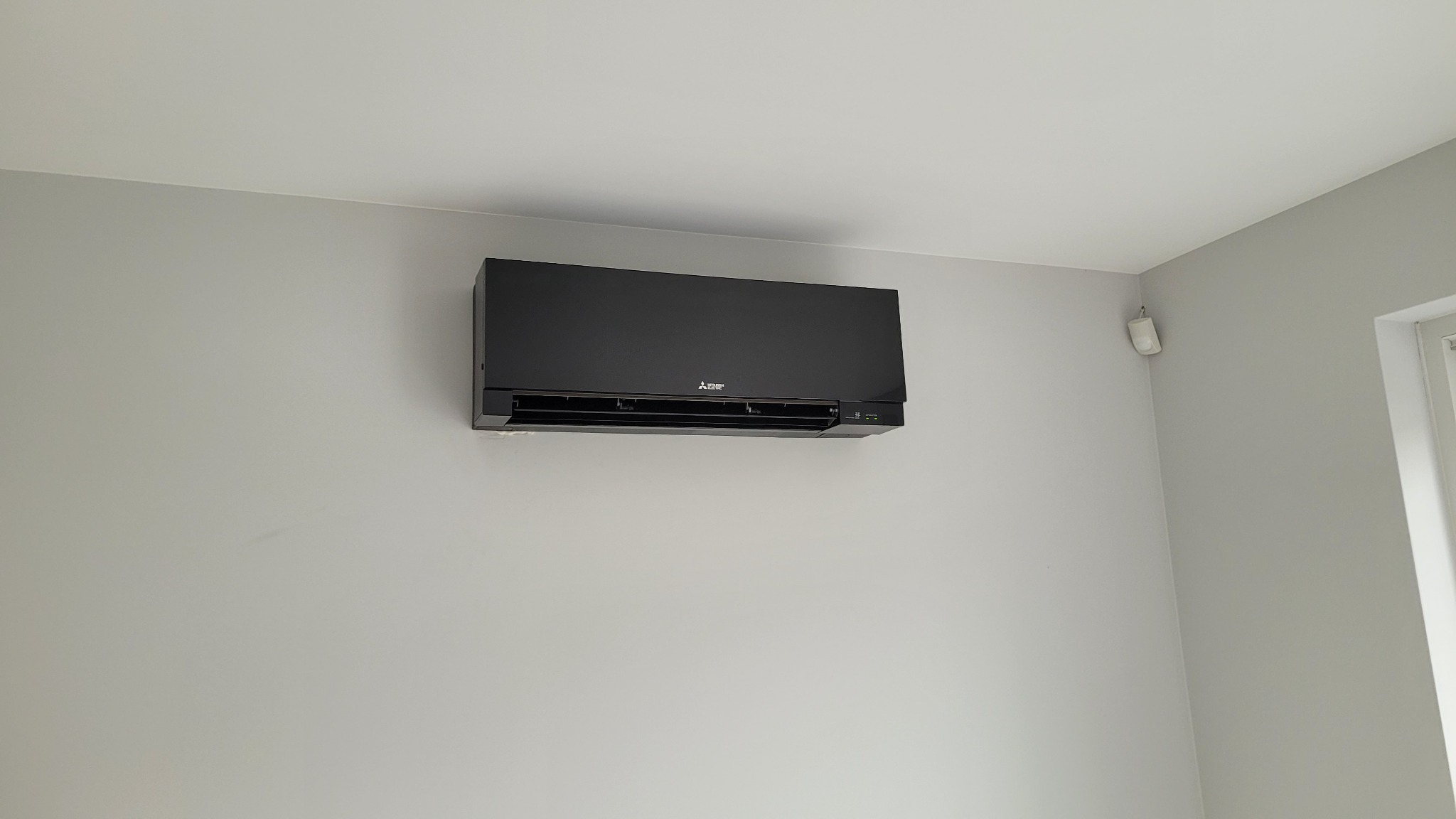 Czarna jednostka klimatyzacji Mitsubishi Electric zamontowana na szarej ścianie w jasnym wnętrzu, widoczny czujnik ruchu na suficie w rogu pokoju.