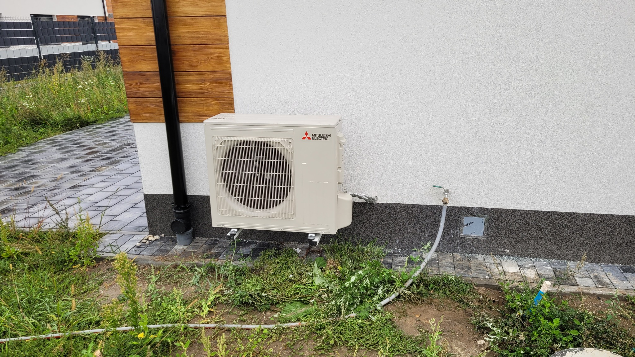 Zewnętrzna jednostka klimatyzacji Mitsubishi Electric zamontowana na elewacji domu, widoczny fragment rynny spustowej i trawnika z kostką brukową.