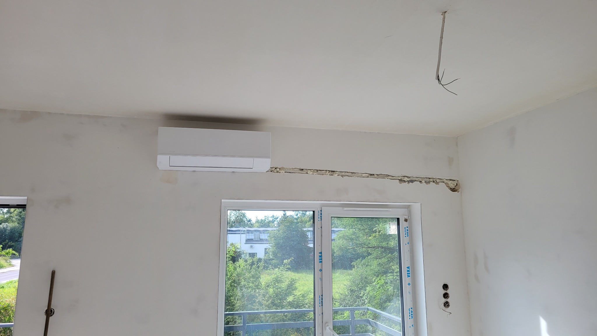 Montaż klimatyzacji w surowym wnętrzu: biały klimatyzator na ścianie nad oknem, widoczne ślady po instalacji i wystające kable z sufitu.