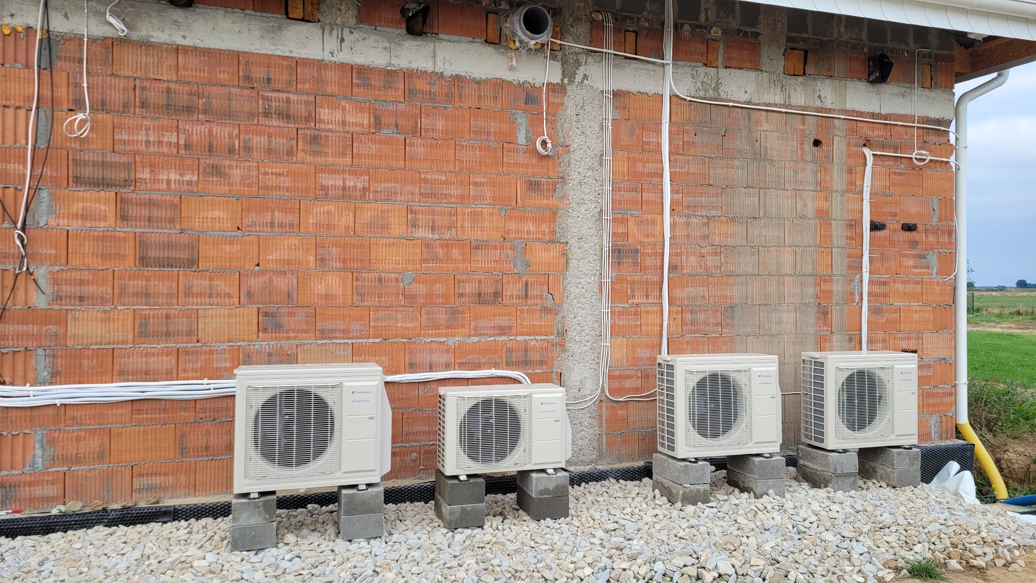 Cztery zewnętrzne jednostki klimatyzacji Fujitsu zamontowane na betonowych blokach przy ceglanej ścianie budynku w trakcie instalacji, widoczne przewody i rury.