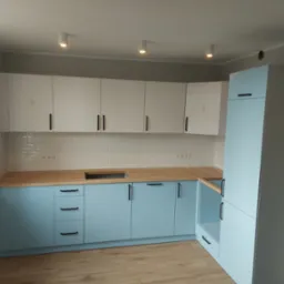 Jasna kuchnia z błękitnymi szafkami dolnymi i białymi górnymi, drewniany blat, białe kafelki na ścianie, minimalistyczne czarne uchwyty.