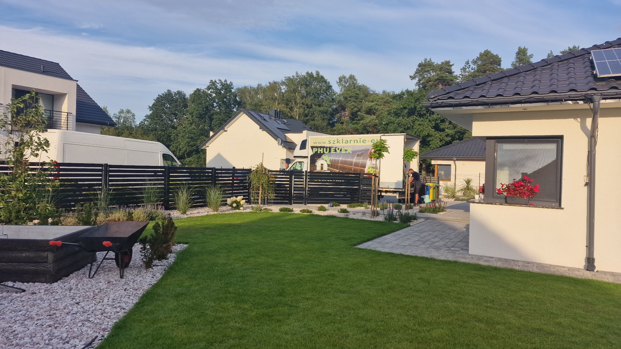 Świeżo założony trawnik z rolki przed nowoczesnym domem jednorodzinnym, widoczna czarna metalowa palisada, biały żwir, taczka i zaparkowany samochód dostawczy z reklamą firmy ogrodniczej.