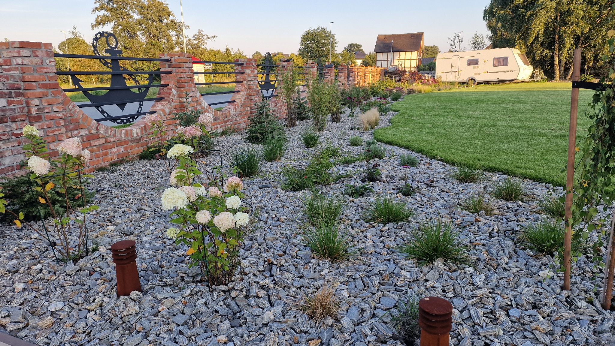 Nowo założony ogród z hydrangeami, ozdobnymi trawami i kamiennym podłożem, ogrodzony ceglanym murem z metalowym ornamentem kotwicy, w tle dom i przyczepa kempingowa.