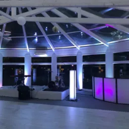 Scena z instrumentami muzycznymi i stanowiskiem DJ-a oświetlona białymi i fioletowymi światłami w przeszklonej sali z drewnianą konstrukcją dachu i lampionami.