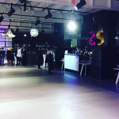 Sala eventowa z parkietem, oświetleniem punktowym, zespołem muzycznym grającym na scenie za podświetlanym DJ-skim stanowiskiem, goście w tle, dekoracyjne lampy wiszące i balony w kształcie cyfr.