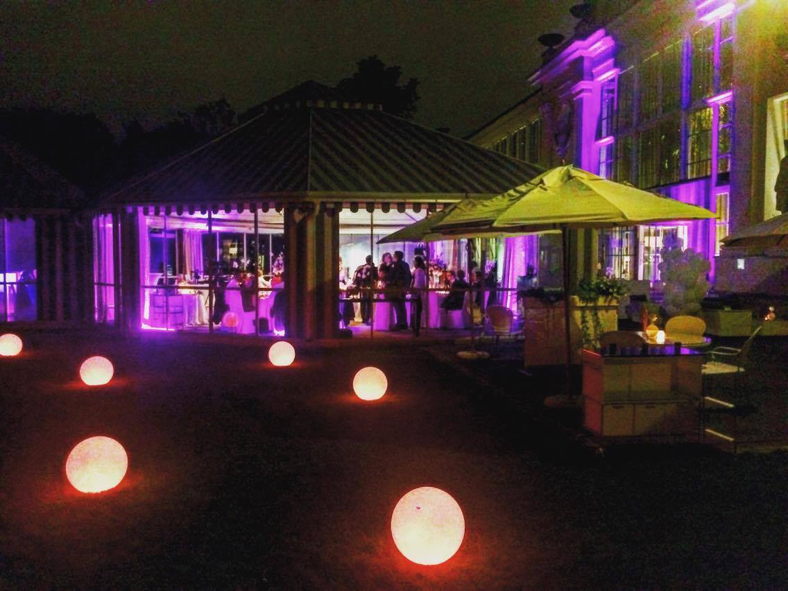 Wieczorny widok na oświetlony ogród z pawilonem, gdzie odbywa się przyjęcie. Dekoracyjne, świecące kule rozproszone na trawniku, fioletowe oświetlenie budynku w tle. Ustawione stoliki i krzesła...