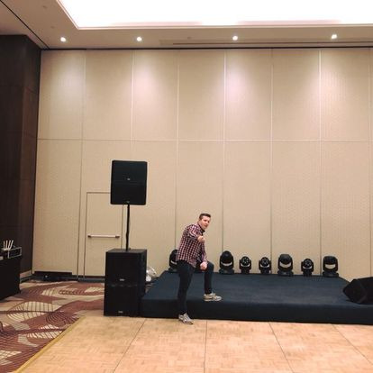 Montaż nagłośnienia i oświetlenia scenicznego: mężczyzna w koszuli w kratę ustawia sprzęt na podwyższeniu, obok kolumny głośnikowe i rząd reflektorów.