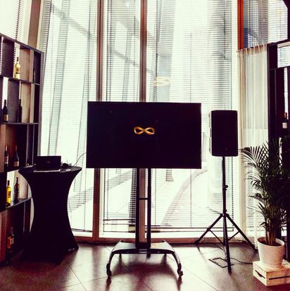 Sala konferencyjna z dużym ekranem wyświetlającym symbol nieskończoności, głośnikiem na statywie, stolikiem koktajlowym i półką z butelkami wina, oświetlona przez okno z żaluzjami.