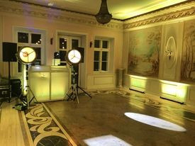 Sala balowa z dekoracyjnym oświetleniem punktowym, konsoletą DJ-ską z podświetleniem i kolumną głośnikową. Na podłodze wzorzysty parkiet. Na ścianie dekoracyjny malunek. Z sufitu zwisa żyrandol.